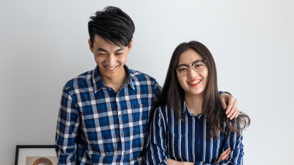 Two people in a well-lit room with plaid and stripped blue shirts smiling at the camera